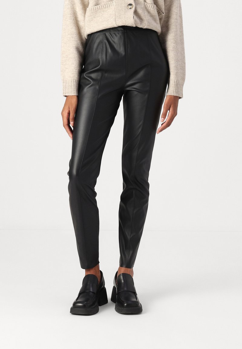 Zwarte faux leren broeken met een strakke uitstraling, hoge taille en opvallende details langs de zijkanten; gecombineerd met zwarte stevige loafers.
