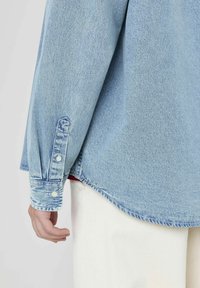 Chemise en denim bleu clair avec un ourlet arrondi, des poignets boutonnés et des détails de couture visibles. Associée à un pantalon en tissu blanc.
