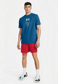 Junger Mann steht, trägt ein blaues Under Armour T-Shirt, rote Shorts, weiße Socken und schwarz-weiße Turnschuhe vor weißem Hintergrund.