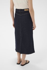 Femme portant une jupe midi en denim bleu foncé taille haute et des sandales noires, debout de dos sur un fond blanc uni.