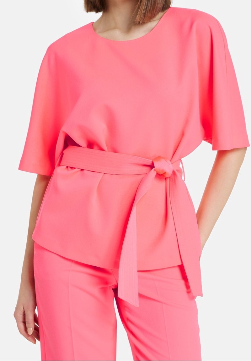 Ljusrosa blus i mjukt tyg, med korta ärmar och ett matchande bälte kring midjan. Slät textur, rundad halsringning.