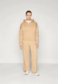 Béžová mikina na zips a zodpovedajúce tepláky z jemnej štruktúrovanej tkaniny. Biely spodný tričko a svetlé topánky dotvárajú vzhľad.