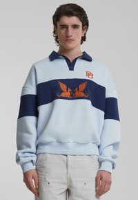 Polo-stil genser i lys blå, med en marineblå stripe, oransje brodert logo, og et grafisk motiv av bevingede skapninger. Blandingsmateriale av bomull.