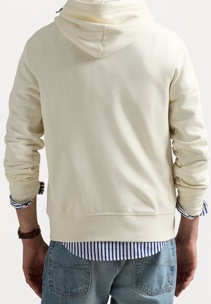Persoon draagt een crèmekleurige hoodie over een blauw-wit gestreept overhemd met opgerolde mouwen en lichtblauwe spijkerbroek, gezien van achteren.