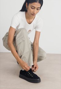 Schwarze Wildledersneaker mit goldenen Ösen und einer dicken schwarzen Sohle. Die Person trägt ein weißes T-Shirt und helle beige Cargo-Hosen.