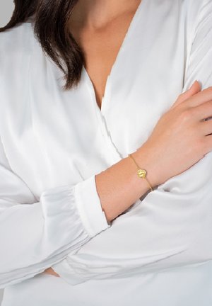 Pulsera de oro con un charm circular, que presenta un acabado suave y una cadena delicada, llevada en una muñeca con una blusa blanca.