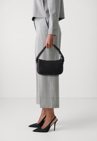 Melnas ādas rokassoma ar pīti rokturi, kuru tur persona pārbaudītā uzvalkā un melnās augstpapēžu kurpēs.