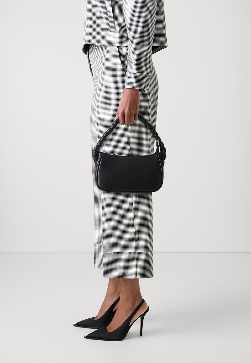 Melnas ādas rokassoma ar pīti rokturi, kuru tur persona pārbaudītā uzvalkā un melnās augstpapēžu kurpēs.