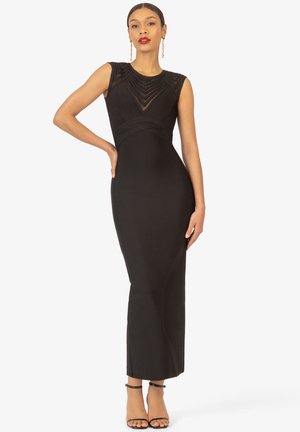 Robe maxi noire avec un design sans manches, corsage texturé et silhouette ajustée. Présente une jupe longue et lisse ainsi que des sandales minimalistes à talons noirs.
