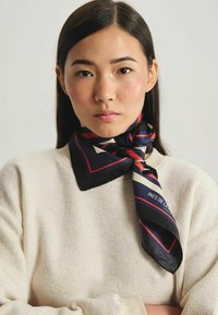 Foulard en soie avec une base marine et des motifs géométriques rouges et crème, noué autour du cou, associé à un pull crémeux texturé.