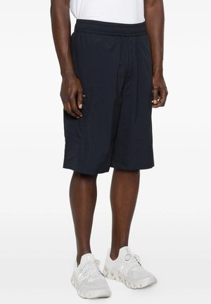 Uomo con pantaloncini neri fino al ginocchio e scarpe da ginnastica bianche in piedi davanti a uno sfondo uniforme.