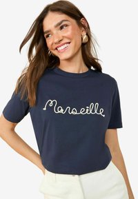 Tamsiai mėlynas medvilninis marškinėlis su balta siuvinėta užrašu "Marseille" ant krūtinės. Trumpomis rankovėmis ir laisvu sijonu.