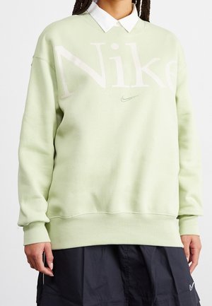Femme portant un sweat-shirt Nike vert clair sur une chemise blanche à col et une jupe noire avec le logo Nike, debout devant un fond uni.