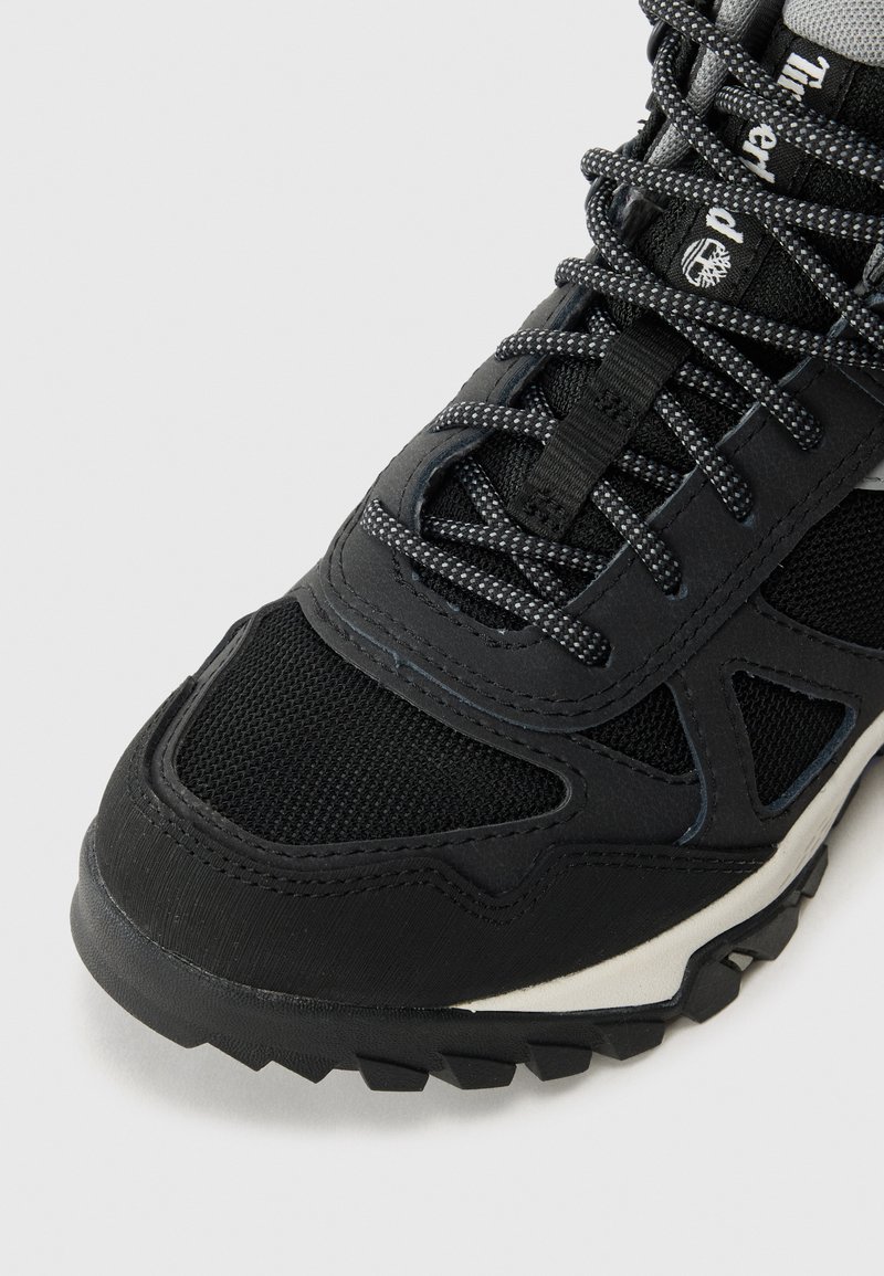 Gros plan sur une chaussure de randonnée noire avec des lacets rayés, des panneaux en mesh et une semelle robuste sur un fond blanc.