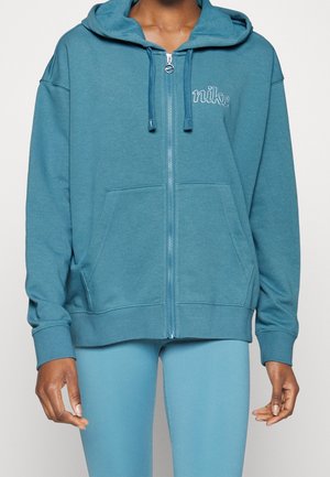 Personne portant un sweat à capuche zippé Nike bleu sarcelle avec poches avant et des leggings bleus assortis, debout devant un fond uni.