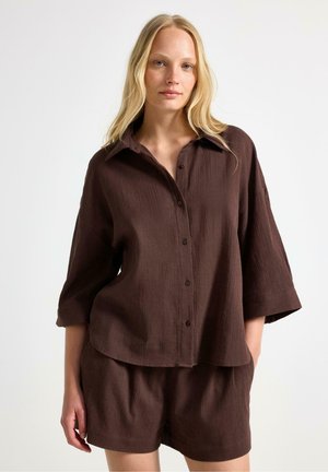 Lyshåret kvinde iført løs brun skjorte med knapper og vide ærmer samt matchende shorts, stående mod en simpel lys baggrund.