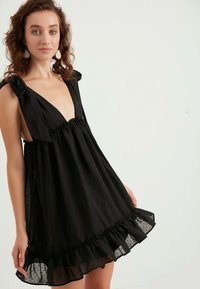Schwarzes ärmelloses Kleid mit tiefem V-Ausschnitt, glattem Stoff, gepunkteter Textur,rüschengesäumtem Saum und Schleifenakzenten an den Schultern.