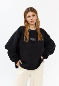 Felpa nera oversize con collo a coste e maniche voluminose. Presenta un logo bianco ricamato sul petto. Abbinata a pantaloni chiari.