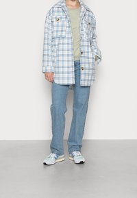 Giacca a camicia a quadri bianca e blu, con grandi tasche e bottoni marroni, abbinata a una maglietta verde chiaro e jeans blu, completata con scarpe da ginnastica.