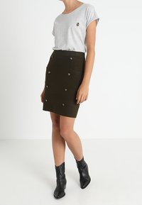 Grå kortärmad T-shirt med bi-broderi, kombinerad med en mörkgrön A-linjeformad kjol med dubbelknappning. Svarta ankelskoot.