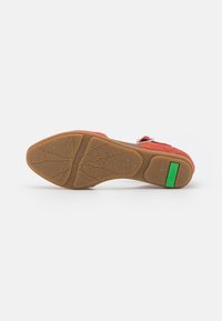 Suela de zapato de ante rojo con fondo de goma texturizada color beige y etiqueta rectangular verde cerca del talón, dispuesta plana sobre fondo blanco.