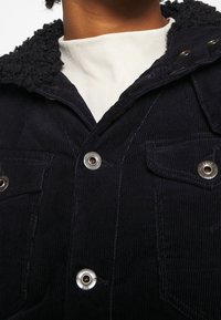 Marinblå kordjacka med en texturerad, fluffig krage, utrustad med metallknappar och dubbla bröstfickor. Närbild av tygdetaljer.