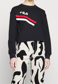 Sweat-shirt FILA noir à manches longues avec des rayures rouges et blanches, associé à un pantalon blanc taille haute avec des motifs noirs abstraits.