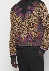 Leopardmönstrad sweatshirt med svarta detaljer, prydd med färgglada paisleymönster och dekorativt hårdvara vid ärmsluten och nederkanten.