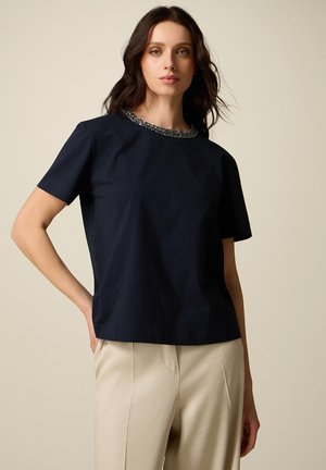 Donna con capelli scuri che indossa una maglietta a maniche corte blu navy con scollo decorato da perline e pantaloni beige a vita alta su sfondo neutro.