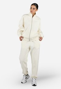 Crème-kleurige zip-up sweatshirt met een staande kraag, gecombineerd met bijpassende tapered sweatpants, beiden gemaakt van een zachte stof. Verrijkt met subtiele branding.