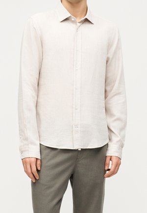Homme portant une chemise beige clair à manches longues boutonnée et un pantalon gris, debout devant un fond blanc uni.
