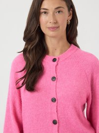 Mulher com cabelo longo e escuro usa um cardigan rosa choque abotoado e brincos de argola dourados, contra um fundo branco.