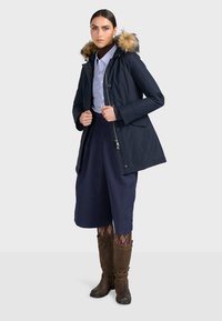 Námornícky modrý parka kabát s kapucňou s umelou kožušinou, zapínaním na zips a postrannými vreckami. Modelka má na sebe košeľu s golierom a hnedé čižmy po kolená.