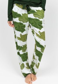 Persoon in witte loungebroek met grote groene bladpatronen en een donkergroen langermige top, staande op blote voeten op een witte ondergrond.