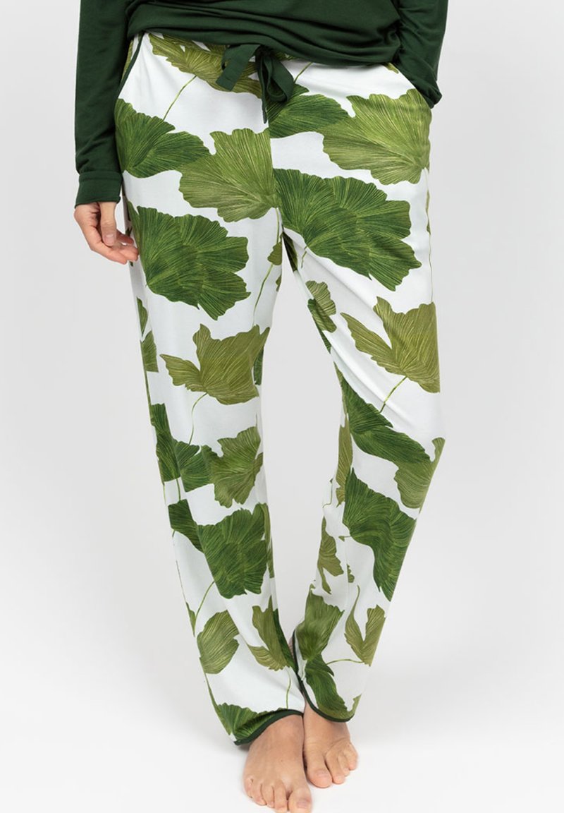 Persoon in witte loungebroek met grote groene bladpatronen en een donkergroen langermige top, staande op blote voeten op een witte ondergrond.