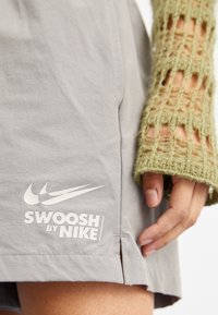Šedé šortky so Swoosh logom a grafikou "SWOOSH by NIKE" na leme. Majú bočné rozparky a hladkú textúru látky.