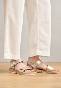 Gold metallic sandals with two straps, cork sole, and contrasting white midsole, featuring a round chain accent on the ankle strap.