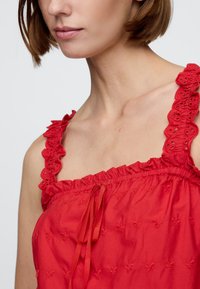 Piros blúz virágos hímzéssel, fodros vállpántokkal és fodros nyakkivágással. Könnyű anyagból készült, elöl húzózsinórral.