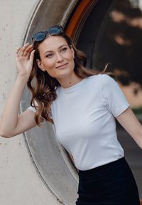 Bílý žebrovaný tričko s krátkými rukávy a kulatým výstřihem, spárované s tmavou sukní. Černé sluneční brýle leží na vlasech. Kulaté okno v pozadí.