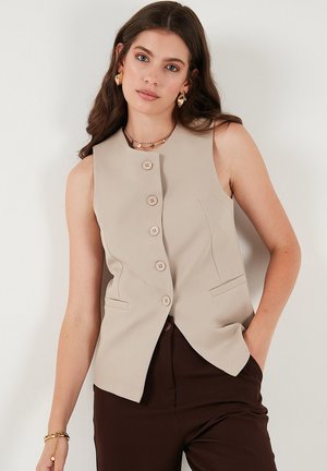 Beige ärmellose Weste mit sechs hellen Knöpfen, seitlichen Taschen und tailliertem Schnitt, aus einem glatten Stoff gefertigt, getragen über dunkelbraunen Hosen.