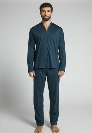 Man with dark hair and beard stands barefoot wearing dark blue button-up long sleeve top and matching pants against light gray background.