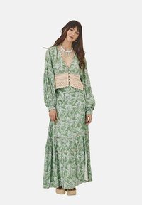 Vestido maxi de patrón floral verde con mangas largas y un cuerpo de ganchillo. Presenta una falda en capas y detalles de botones. Se lleva con zapatos de plataforma.
