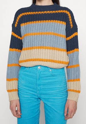 Osoba ubrana w sweter w paski w kolorach granatowym, jasnoniebieskim, beżowym i pomarańczowym, zestawiony z jasnoniebieskimi spodniami sztruksowymi.