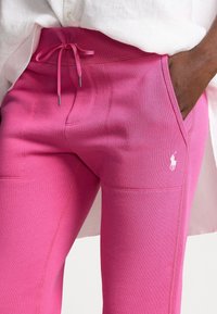 Rosa träningsbyxor med dragsko i midjan, med sidofickor och en liten vit logotyp. Mjuk textur och åtsittande stil.