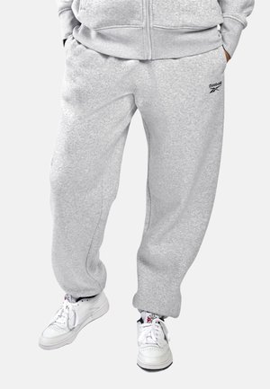 Persoon die lichtgrijze Reebok joggingbroek draagt, bijpassend vest met ritssluiting, witte Reebok sneakers en zwarte sokken met een rood-wit logo.