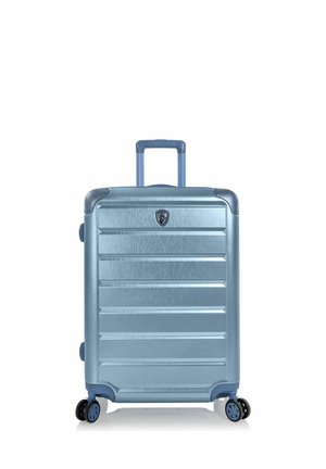 Valigia rigida azzurro chiaro con manico telescopico e quattro ruote doppie girevoli, in piedi su sfondo bianco.