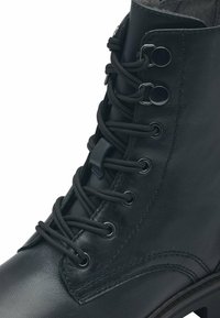 Bota de cuero negro hasta el tobillo con punta redonda, superficie texturizada, cinco pares de ojales y cordones negros. Presenta una suela gruesa y detalles de costura.
