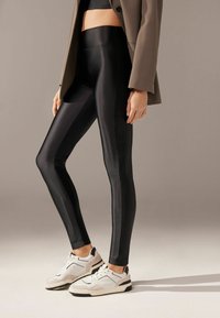 Leggings negros y brillantes hechos de un material suave y elástico, combinados con zapatillas blancas y un blazer neutro, que muestran una silueta ajustada.