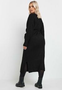 Musta neulejakku mekko, jossa on vyötärösolki, pitkät hihat, sivuhalkiot ja puolipitkä pituus, yhdistettynä paksuihin mustiin nilkkureihin.