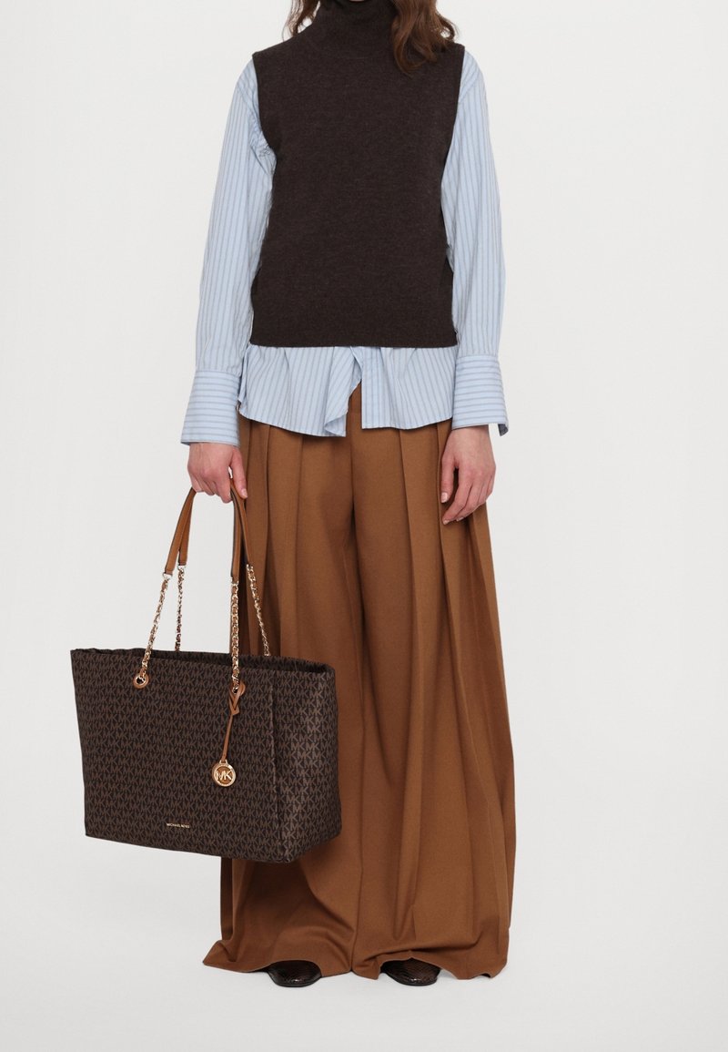 Personne portant un pull sans manches marron foncé par-dessus une chemise à rayures bleues et un pantalon large marron, tenant un sac fourre-tout marron à motifs.